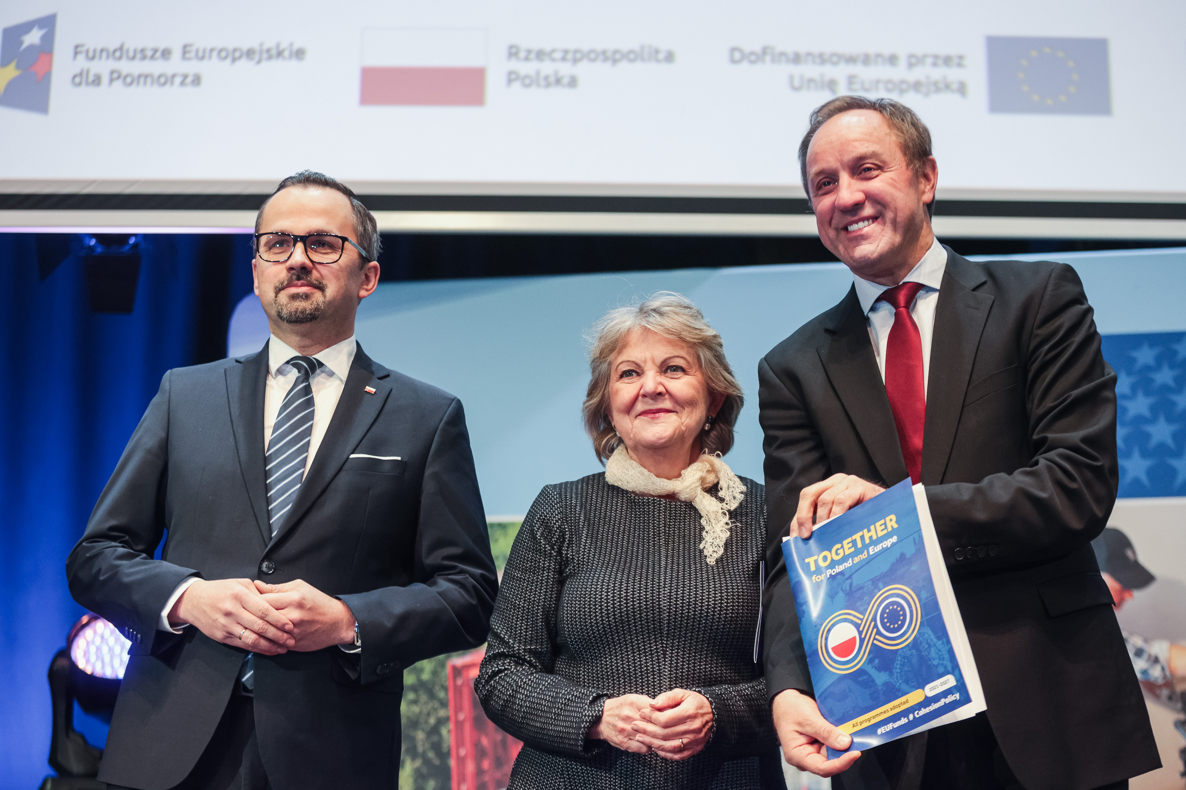  Zdjęcie z inauguracji otwarcia programu Fundusze Europejskie dla Pomorza 2021-2027 - FEP. Przedstawia ono trzy osoby. Od lewej sekretarz stanu w Ministerstwie Funduszy i Polityki Regionalnej Marcin Horała, obok niego, po środku, Unijna komisarz Elisa Ferreira, a po prawej jest marszałek Województwa Pomorskiego Mieczysław Struk. Marszałek trzyma decyzję osobiście przekazaną przez panią komisarz (fot. pomorskie.eu).